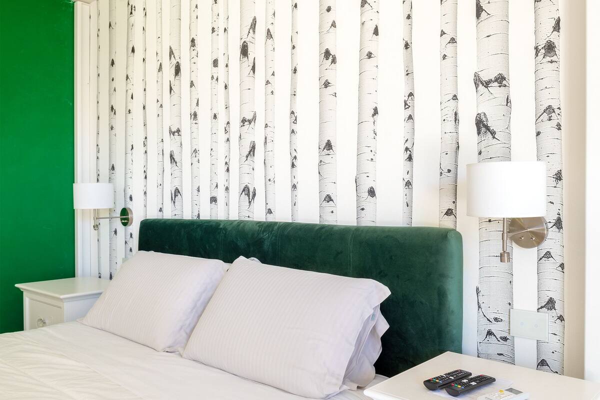 A neatly made bed with white linens, a green headboard, and a nightstand with a lamp and remote controls.