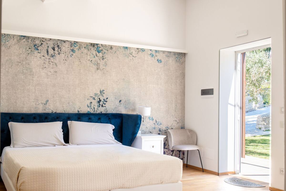 A bedroom with a large bed, a nightstand, a chair, and a view of the outdoors.