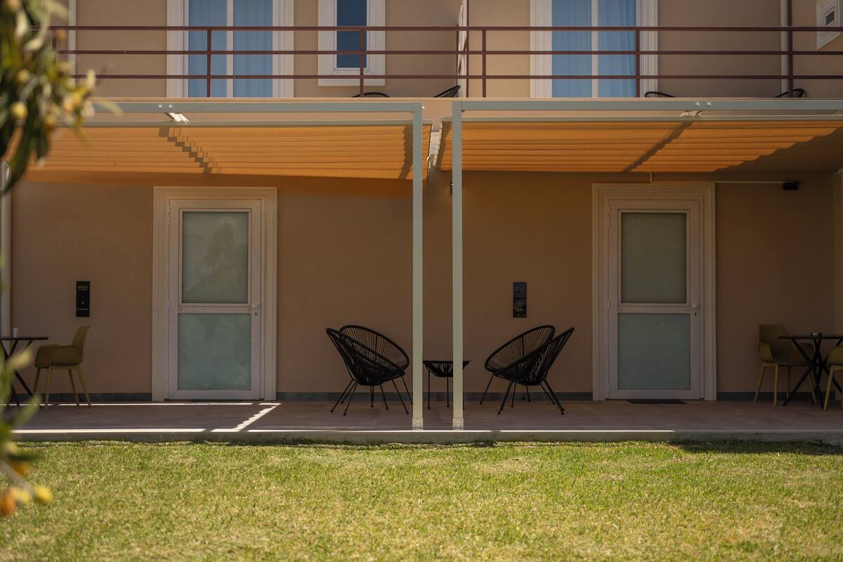 A building with two doors, a covered outdoor seating area with chairs and tables, and a grassy lawn in front.
