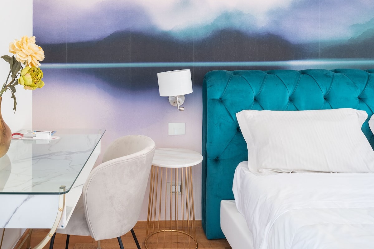 A bedroom with a bed, a glass table, a chair, a lamp, and a painting of a landscape.
