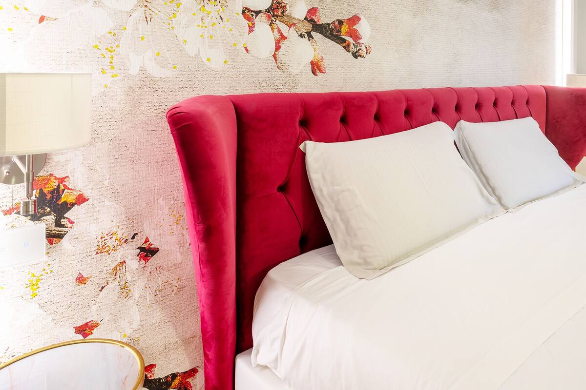A bedroom with a red upholstered headboard, white bedding, and a textured wall with floral patterns.