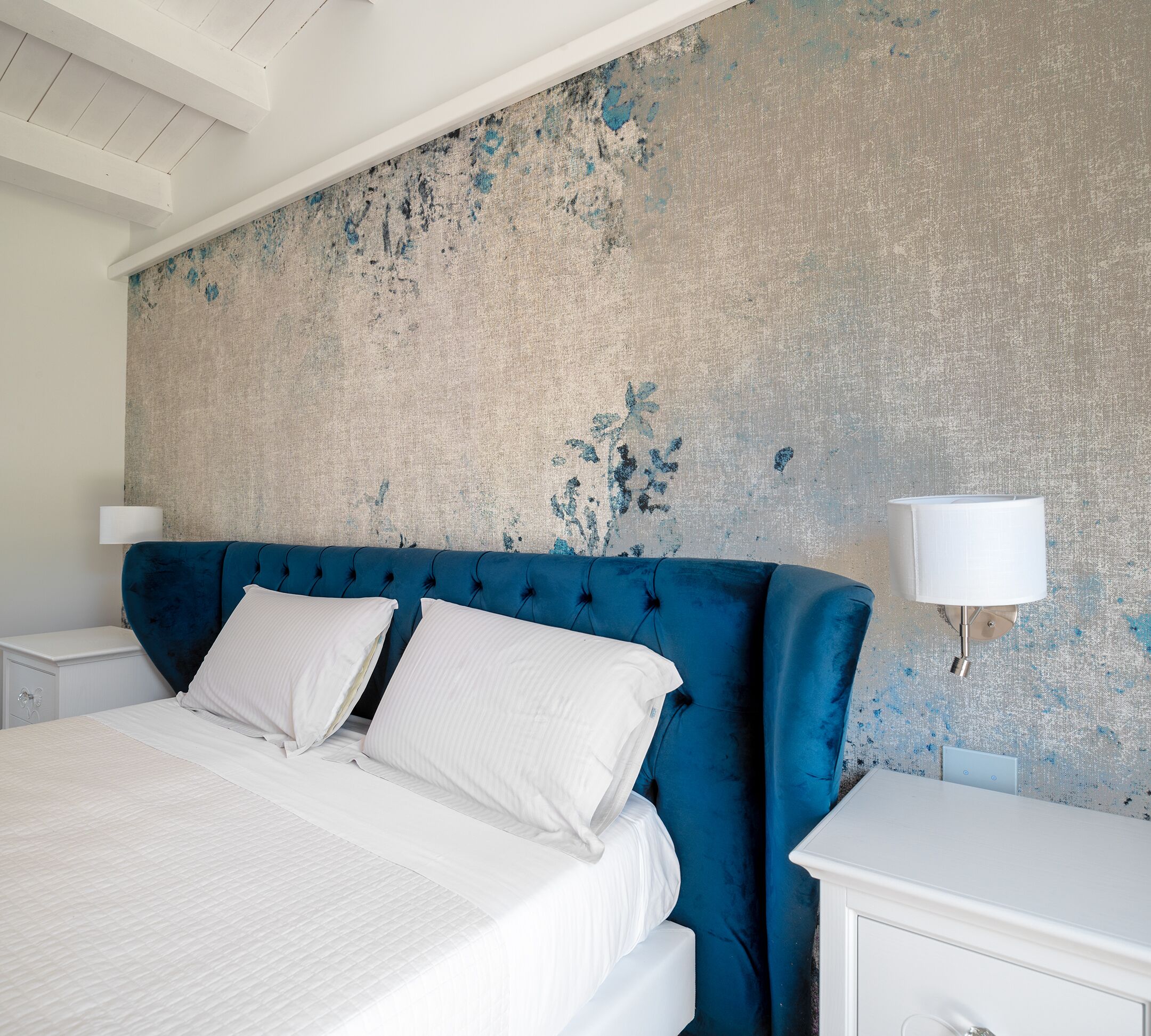A bedroom with a large bed, a blue upholstered headboard, white bedding, and a textured wall.