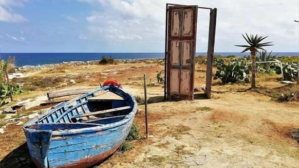 Un bote azul sobre un sendero de tierra con una puerta de madera y una palmera al fondo.