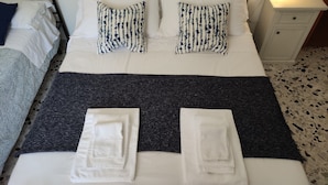 Una cama bien hecha con almohadas blancas y estampadas, un edredón oscuro y toallas dobladas.