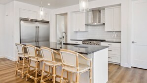 A modern kitchen with white cabinetry, a stainless steel refrigerator, a central island with bar stools, and pendant lighting. - 1 Chill Casa: Desert Color Lagoon + Beaches (St George)