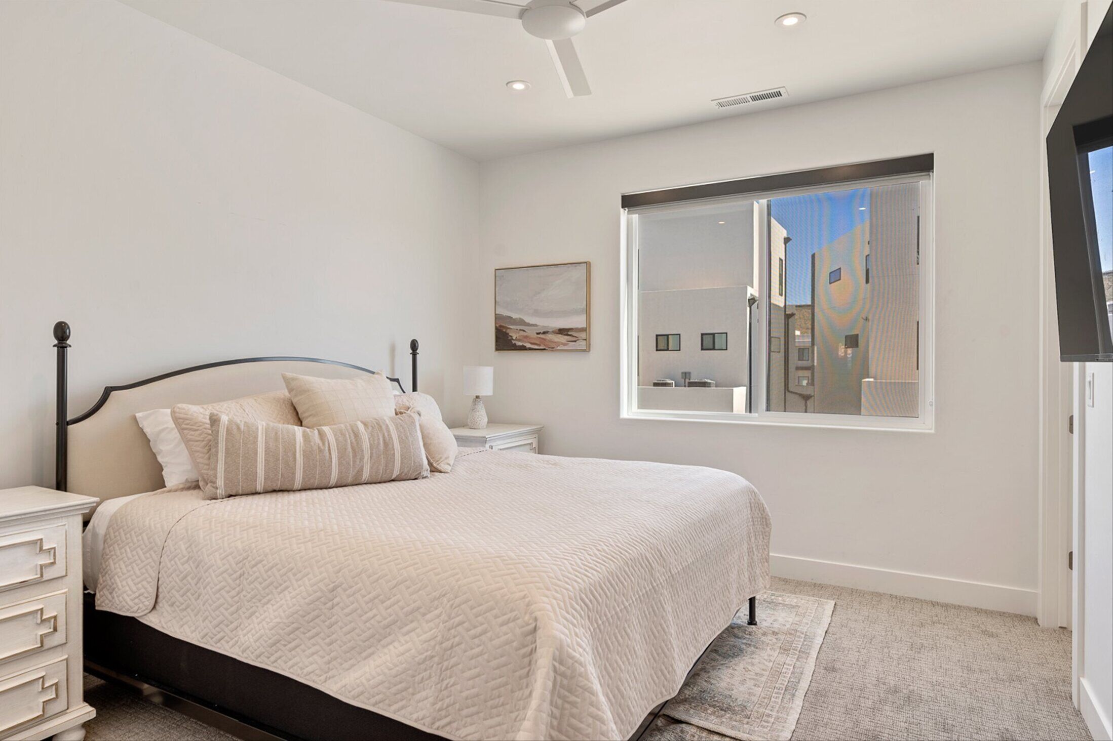 A bedroom with a bed, nightstands, a lamp, a window with a view of buildings, and a ceiling fan.