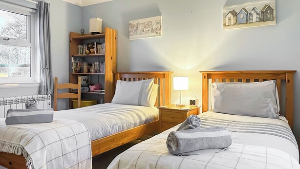 A bedroom with two wooden beds, a bedside table with a lamp, a bookshelf, and a window with curtains. - 2 bedroom accommodation in Bamburgh (Bamburgh)