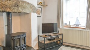 Ein Wohnzimmer mit Kamin aus Stein, einem Fernseher auf einem Holzboden und Vorhängen vor einem Fenster. Ein Heizkörper ist ebenfalls zu sehen.