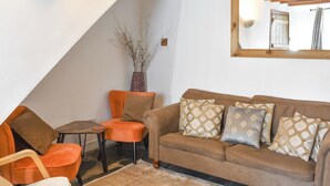 Ein Wohnzimmer mit einem braunen Sofa, einem orangefarbenen Sessel und einem kleinen Tisch. An einer weißen Wand steht eine Vase mit Trockenpflanzen.