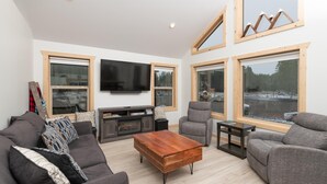 A modern living room with a large flat-screen TV, grey sofas, a wooden coffee table, and large windows offering a view of a snowy landscape. - The Alpine Cabin Rental (British Columbia)
