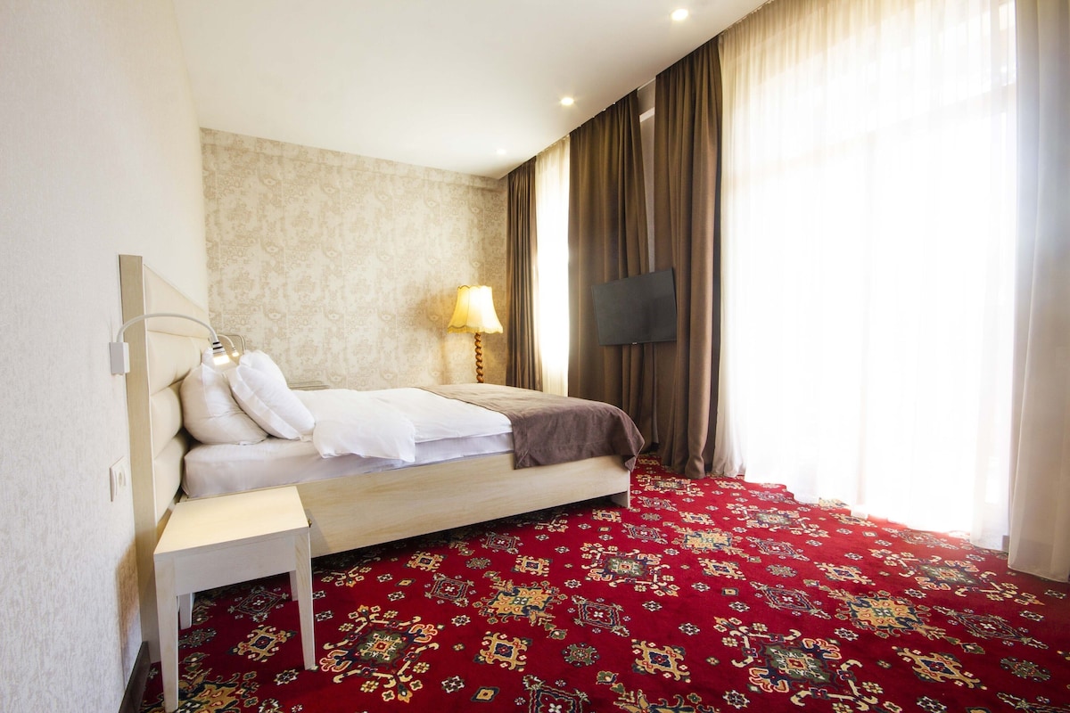 A hotel room with a bed, a bedside table, a lamp, a TV, and a patterned carpet.