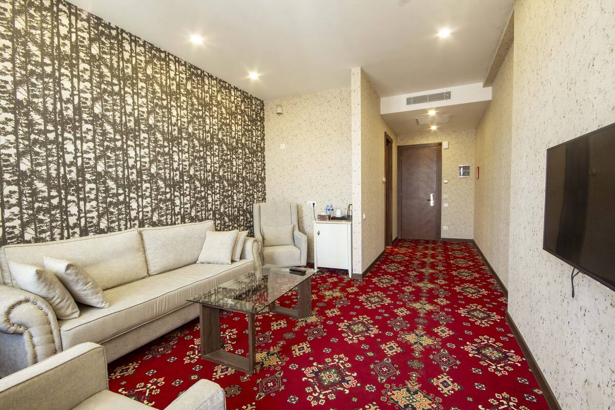 A room with a patterned wallpaper, a sofa, a glass coffee table, a red carpet, and a TV mounted on the wall.