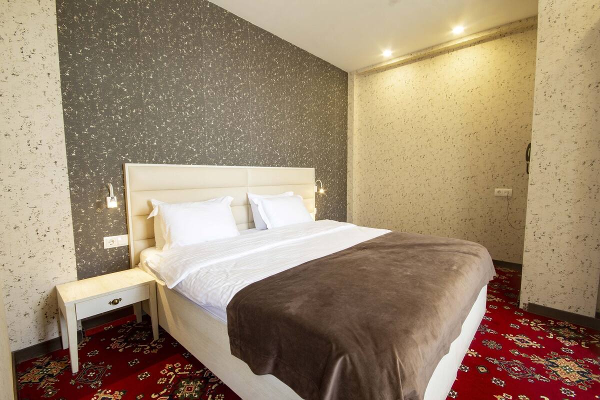 A hotel room with a bed, a nightstand, a patterned carpet, and a textured wall.