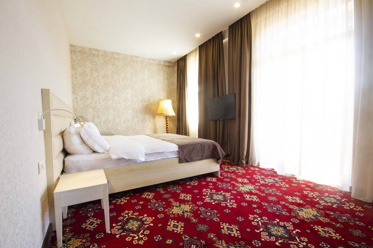 A hotel room with a bed, a bedside table, a lamp, a TV, and a patterned carpet.