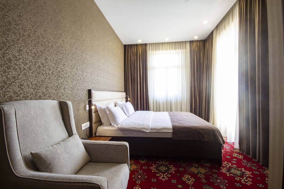 A hotel room with a bed, a chair, a patterned wall, and a red carpet.