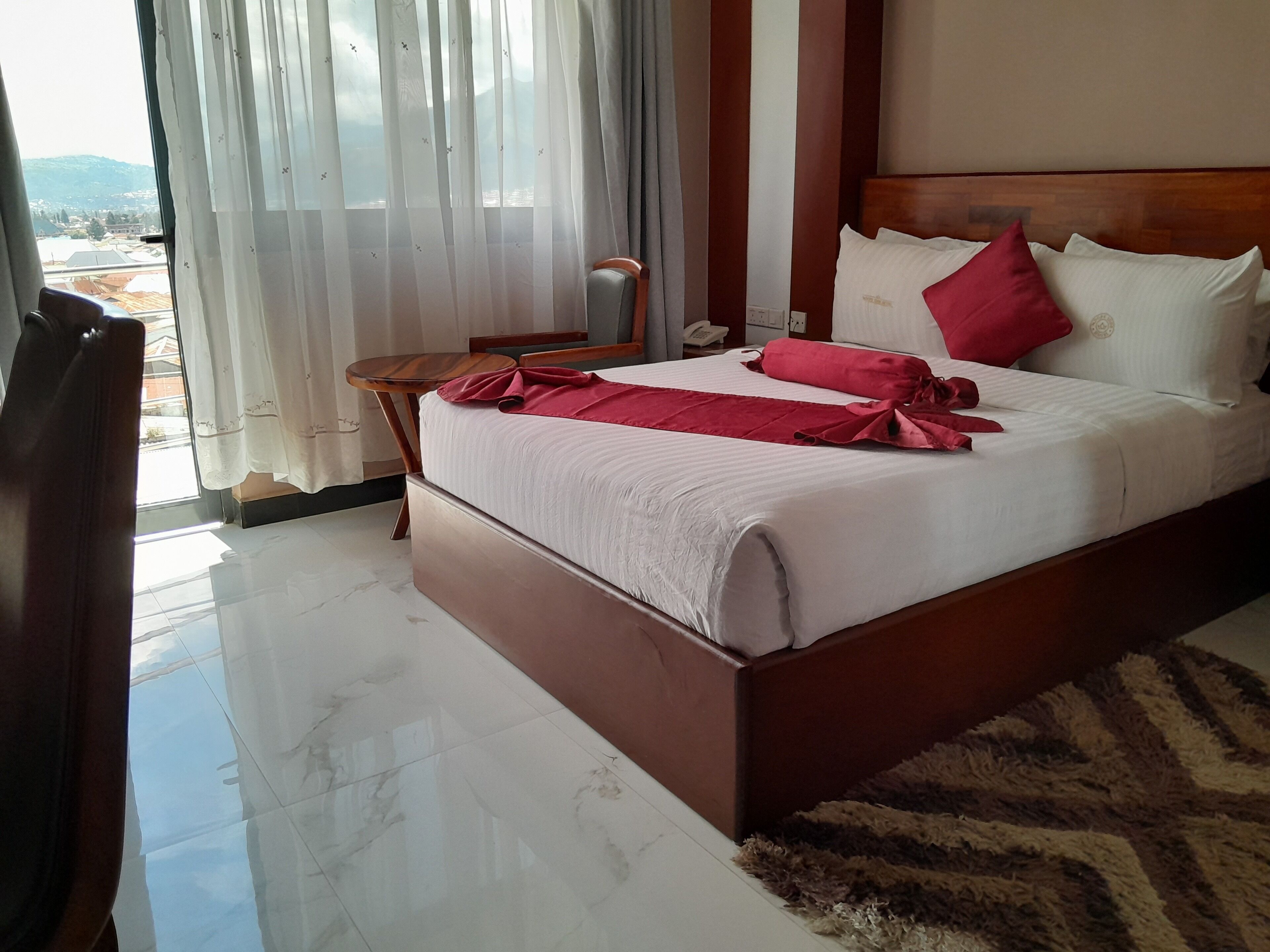 A hotel room with a large bed, a wooden headboard, a small wooden table, a telephone, and a view of the outdoors through sheer curtains.