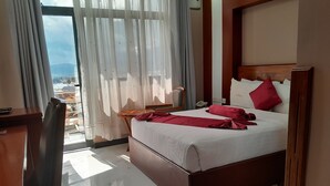 Quarto de hotel com cama, escrivaninha, cadeira, janela com cortinas e vista para edifícios.
