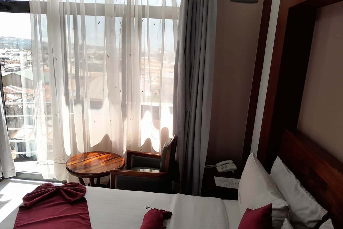 Habitación de hotel con cama, una mesita, una silla y vistas de la ciudad a través de cortinas translúcidas.