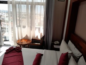 A hotel room with a bed, a small table, a chair, and a view of the city through sheer curtains. - Mdope Idde hotel (Mbeya)