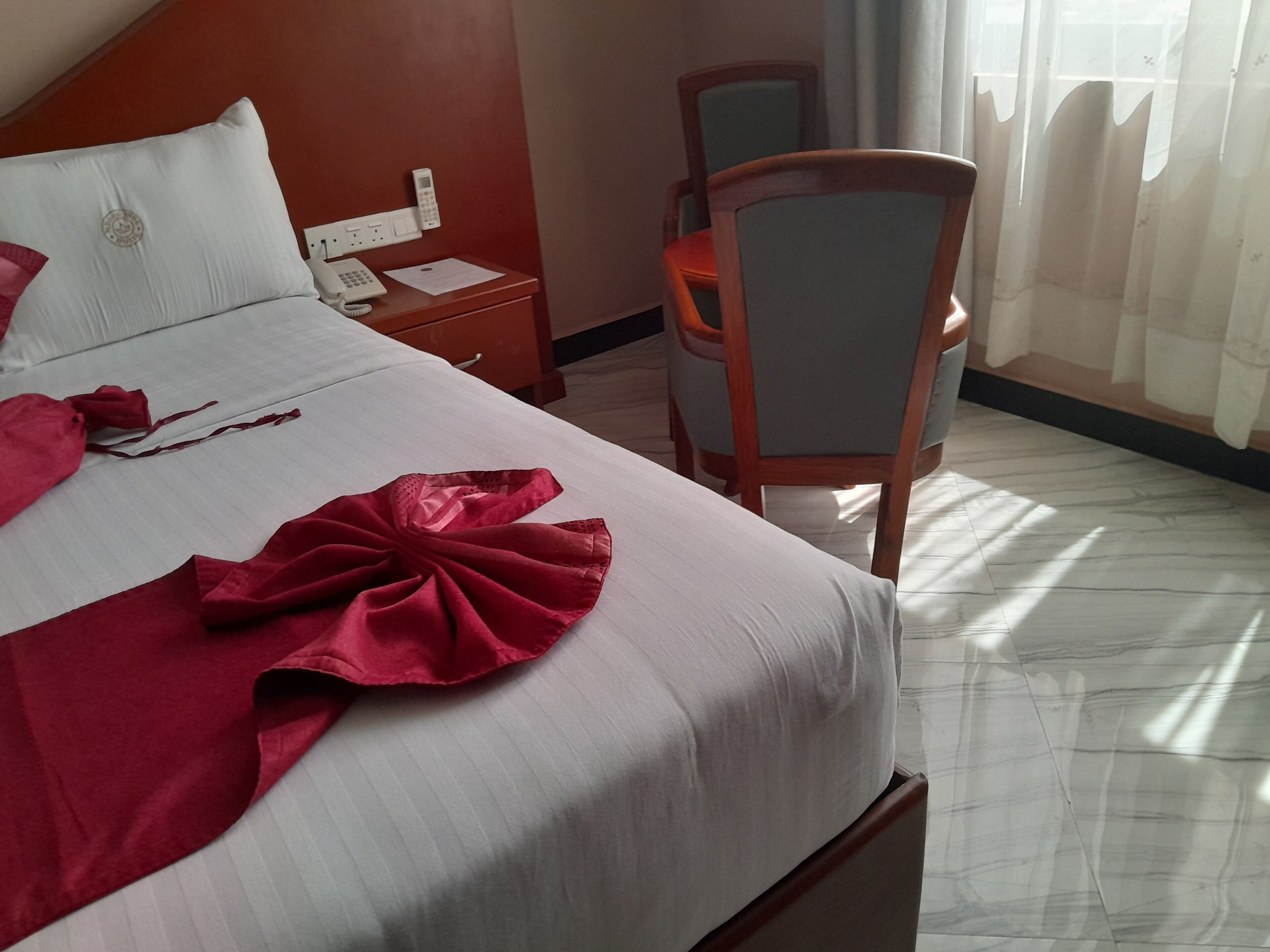 A hotel room with a neatly made bed, a wooden chair, a bedside table with a phone and papers, and a window with curtains.