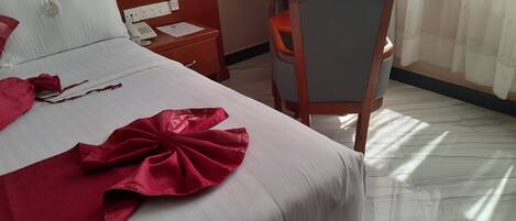 A hotel room with a neatly made bed, a wooden chair, a bedside table with a phone and papers, and a window with curtains.