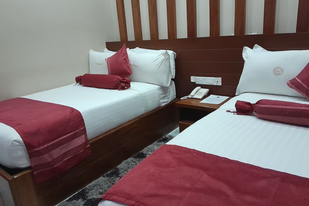 Habitación de hotel con dos camas, cada una con ropa blanca y detalles rojos, un cabecero de madera, una mesita de noche con un teléfono y una libreta, y una alfombra estampada.