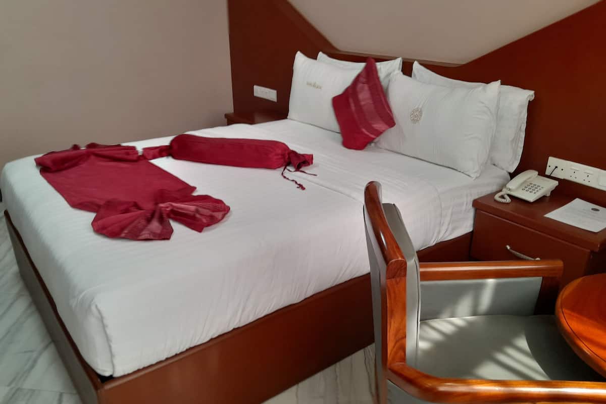 Una cama bien hecha con sábanas blancas y una manta roja, cabecera de madera, una mesita de noche con un teléfono y papeles, y una silla de madera con cojín.