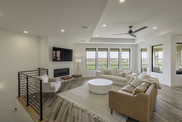 A spacious living room with a large window, a ceiling fan, and a flat-screen TV mounted on the wall. - New 5BR Sleeps 18 - Snow Canyon Views & Hot Tub (St. George)
