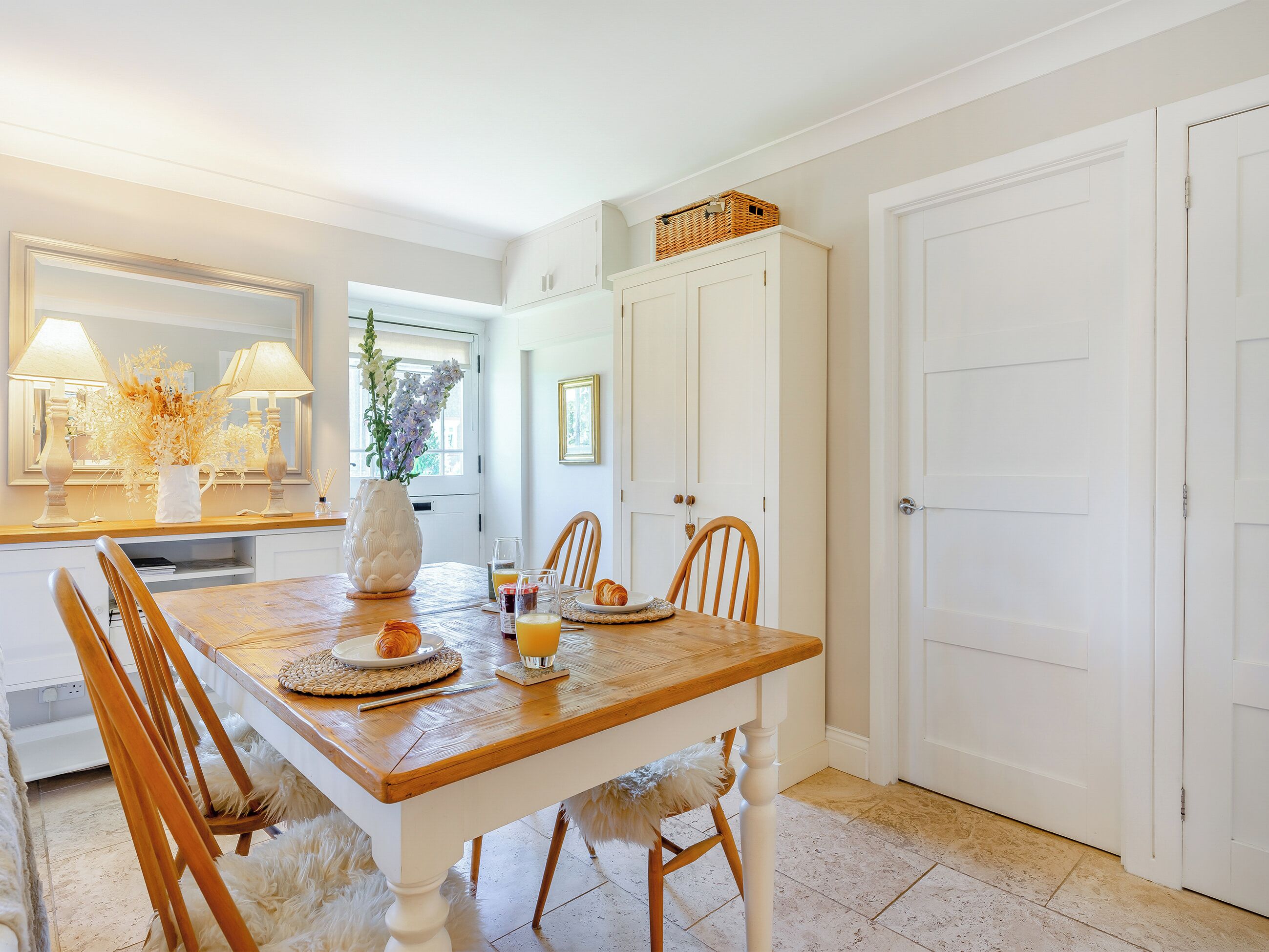 Un comedor con una mesa de madera, sillas y un jarrón de flores. Se observa un mueble blanco sobre la mesa y una puerta a la derecha.