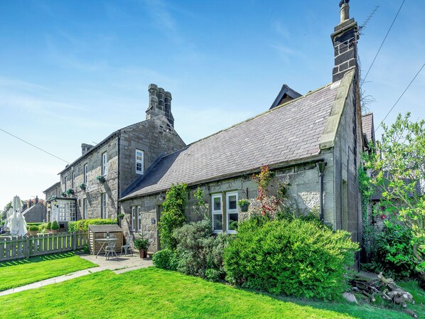 A stone building with a sloped roof, a chimney, and a small garden area with a lawn and potted plants.