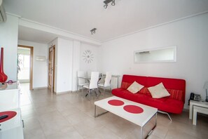 A modern living room with a red sofa, a dining area with white chairs, and a kitchen area with a red guitar. - Two bedroom  in Spain, La Torre Golf Resort (Torre-Pacheco)