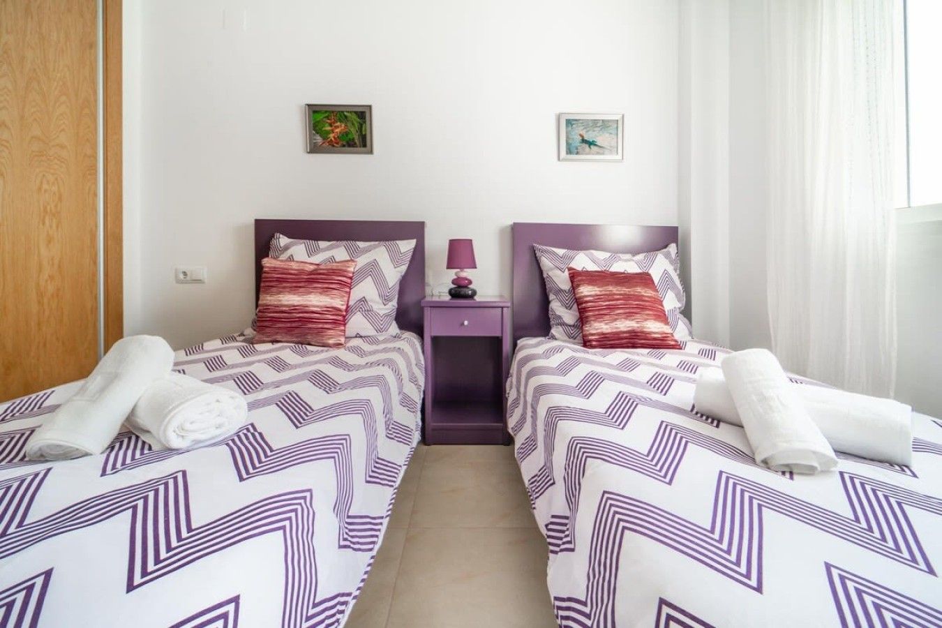 A hotel room with two beds, each with a patterned bedspread and pillows, a nightstand with a lamp, and a small wall art piece above the bed.