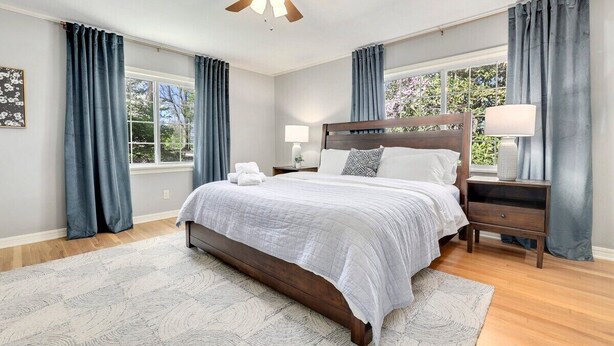 Une chambre à coucher comprenant un lit, des tables de chevet, un ventilateur de plafond et de grandes fenêtres avec des rideaux.