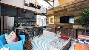 Une cuisine moderne avec un plafond haut, un coin repas avec des tabourets de bar, un salon avec un canapé bleu et un téléviseur fixé à un mur en bois.