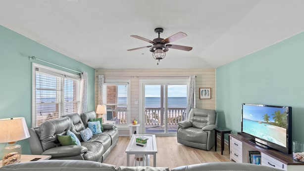 A living room with a large TV, leather couches, a ceiling fan, and a view of the ocean through the sliding glass doors. - Updated 3 BR Oceanfront Condo - Drayton House 402 (Myrtle Beach)