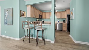 A kitchen with wooden cabinets, a bar stool, and a refrigerator. - Updated 3 BR Oceanfront Condo - Drayton House 402 (Myrtle Beach)