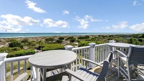 Une terrasse avec une table ronde et des chaises, donnant sur une plage à la végétation clairsemée et sous un ciel dégagé.