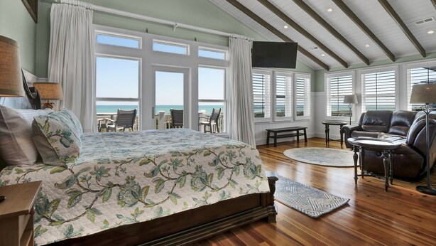 Une chambre spacieuse avec un grand lit, un plancher en bois, une vue sur l’océan et un coin salon comprenant un canapé et des fauteuils.