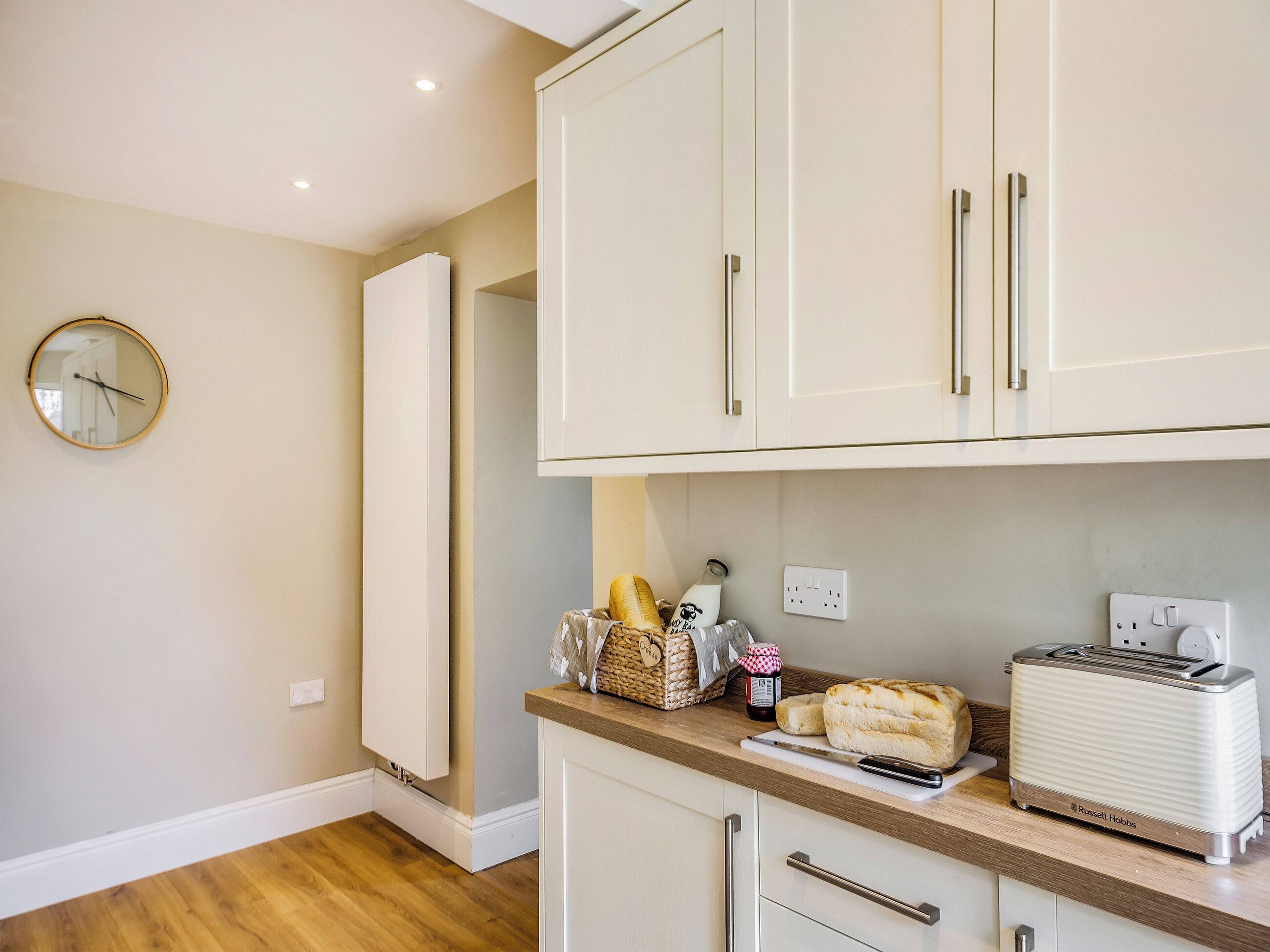 A kitchen with white cabinets, a wooden countertop, a toaster, a bread loaf, a jar of jam, and a clock on the wall.