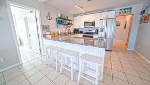 Et køkken med en central ø, hvide barstole og en mikrobølgeovn.