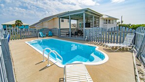 En swimmingpool med træterrasse, liggestole og en overdækket patio.