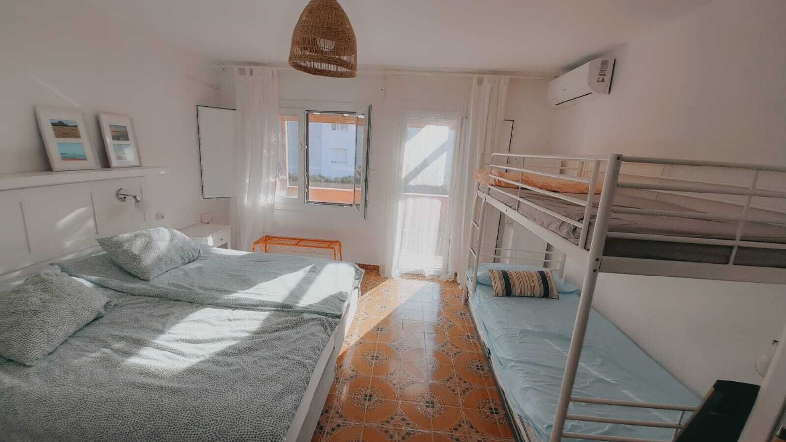 Une chambre de dortoir avec des lits superposés, une fenêtre avec des rideaux, un luminaire suspendu et un sol carrelé.