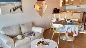 A living room with a beige sofa, a round coffee table, and a dining area with a table covered in a leaf-patterned tablecloth. - Appart vue Mer, 100m de la Plage, Avec Terrasse et Parking Privé (Perros Guirec)