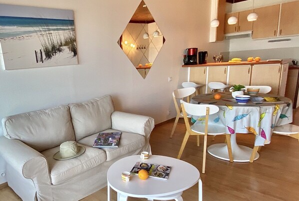 A living room with a beige sofa, a round coffee table, and a dining area with a table covered in a leaf-patterned tablecloth. - Appart vue Mer, 100m de la Plage, Avec Terrasse et Parking Privé (Perros Guirec)