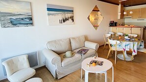 A living room with a beige sofa, a small round table, and a dining table with chairs. There are two paintings on the wall, a kitchen area with cabinets and a countertop, and a wooden ceiling. - Appart vue Mer, 100m de la Plage, Avec Terrasse et Parking Privé (Perros Guirec)