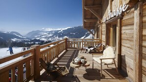 Una terraza de madera con vistas a montañas nevadas, un letrero que dice "Le Chasse" y mobiliario exterior con una mesa y sillas.