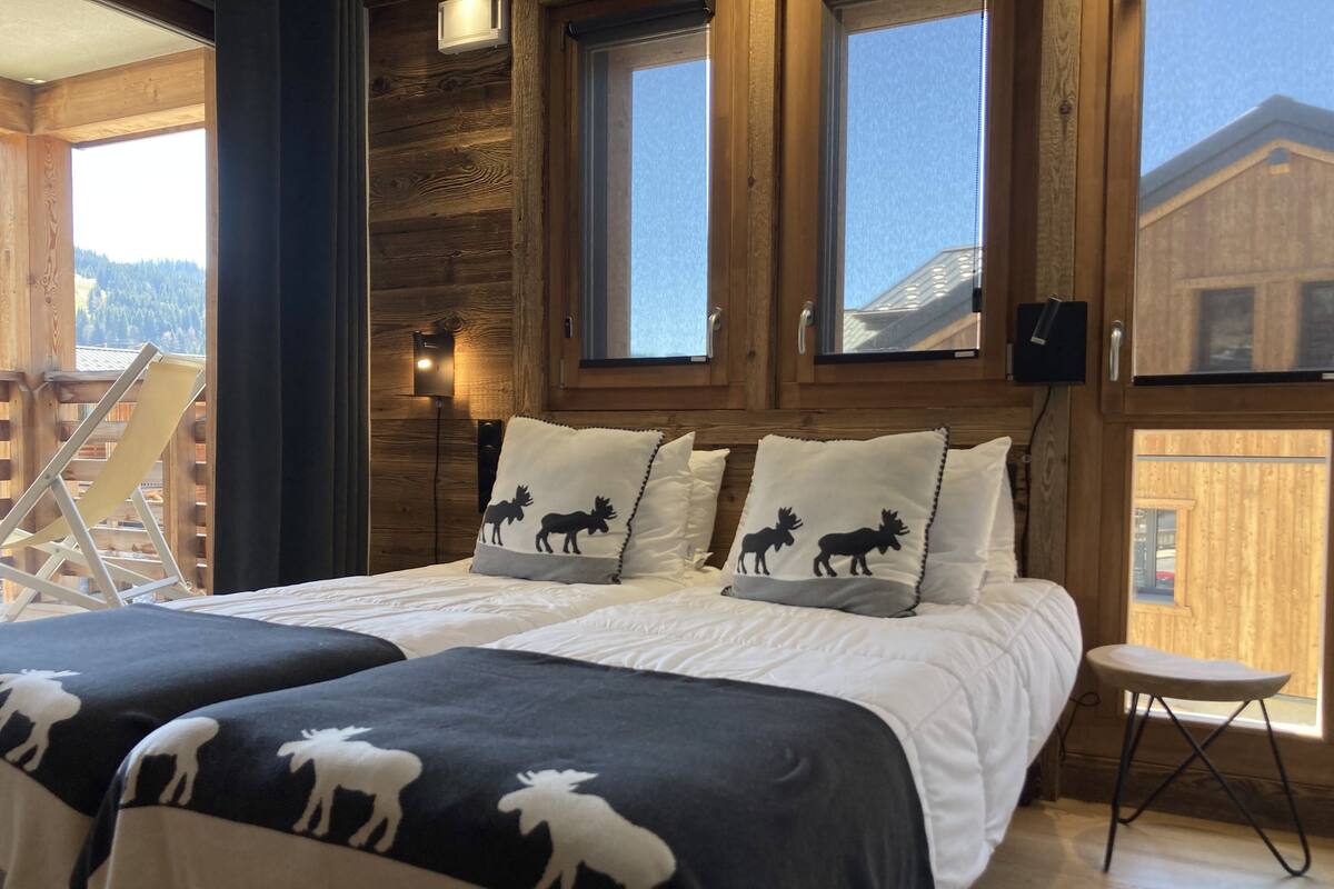 Una habitación con dos camas, cada una con ropa de cama temática de alce, paredes de madera y ventanas amplias que ofrecen vistas al exterior.
