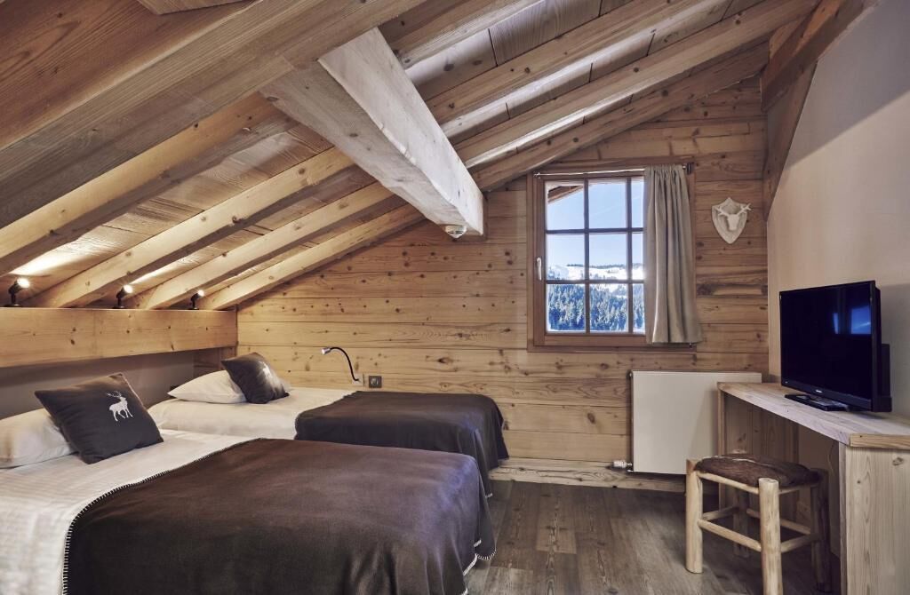A cozy attic bedroom with two beds, a wooden ceiling, a window with a view, and a TV.