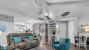 Una sala de estar moderna con un sofá de cuero gris, sillones verde azulado y una alfombra estampada. Una escalera espiral conduce a un nivel superior.