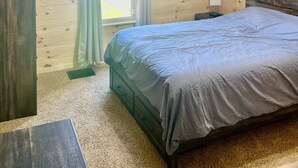 A bedroom with a wooden floor, a bed with a grey bedspread, a ceiling fan, a window with curtains, and a cross on the wall.